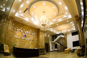 Vestíbulo de hotel con mostrador de recepción, candelabro, escalera y zona de estar.