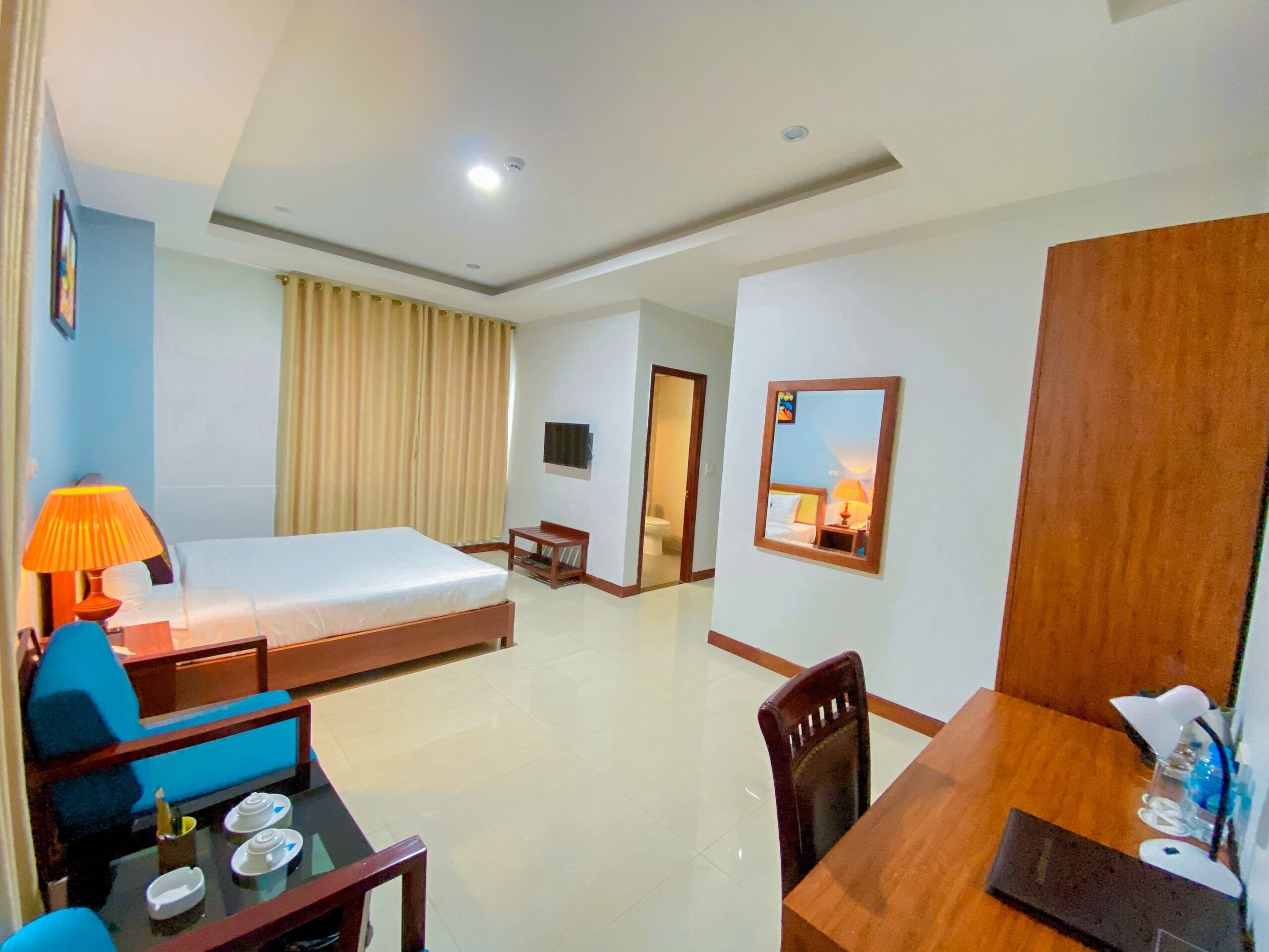 A hotel room with a bed, a desk, a chair, a table with cups, a lamp, and a mirror.