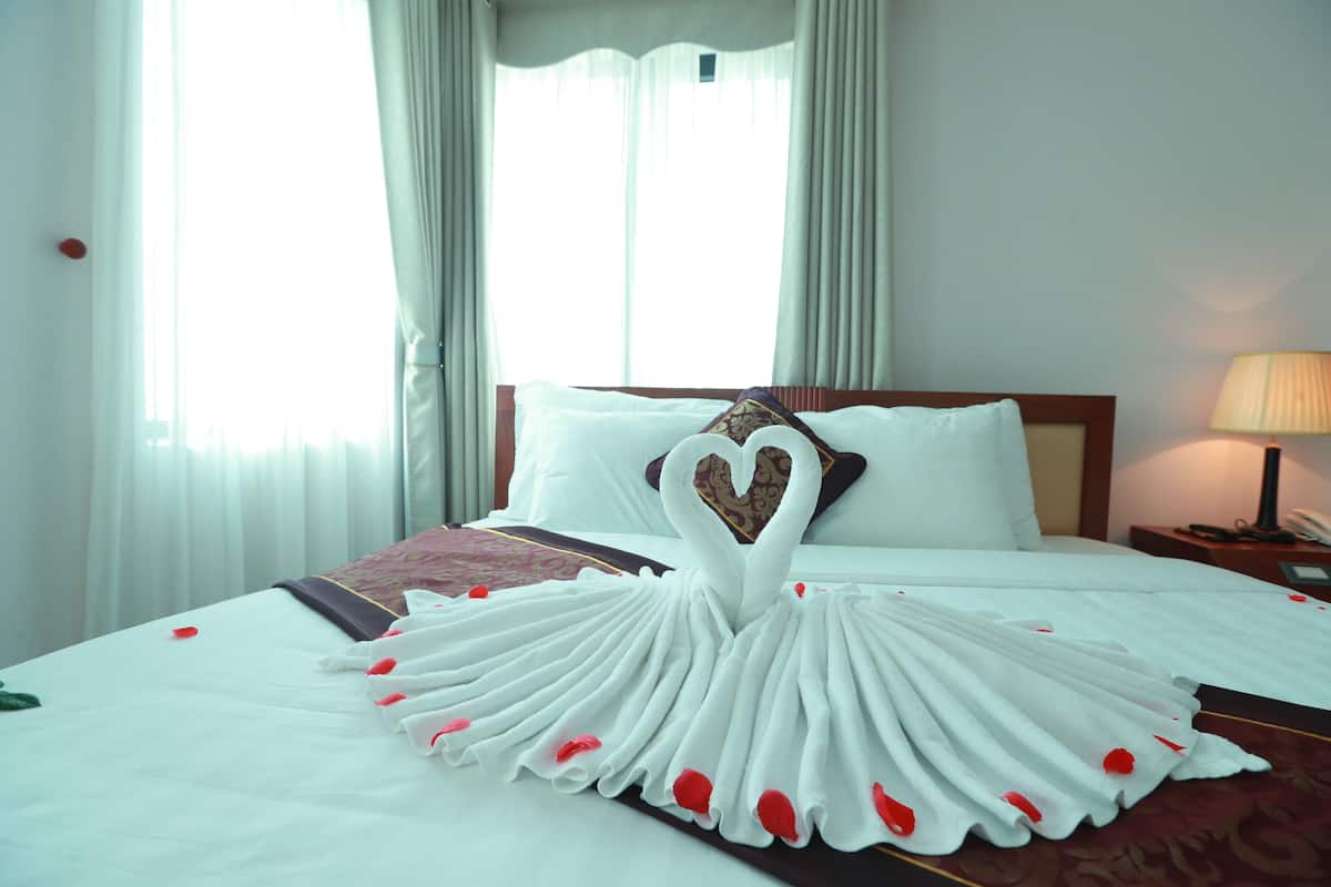 Habitación de hotel con una cama adornada con una toalla en forma de corazón y pétalos de rosa.