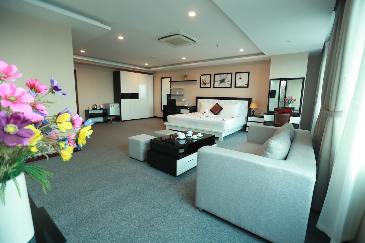 Una habitación de hotel moderna con una cama grande, un sofá, una mesa de centro y un escritorio. Hay un arreglo floral a la izquierda y un espejo a la derecha.