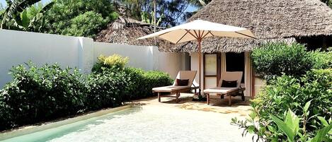 A clear pool with lounge chairs and a thatched-roof structure, surrounded by lush greenery.
