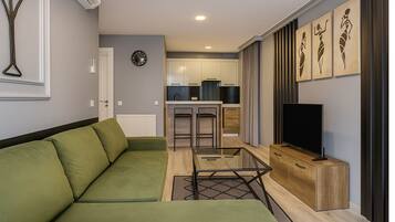 A modern living room with a green sofa, a glass coffee table, a TV on a wooden stand, and a kitchen in the background.