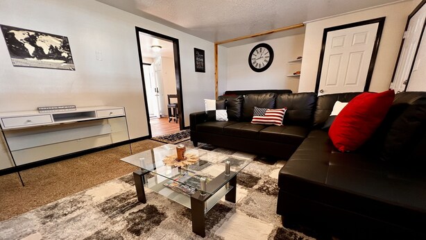 A living room with a glass coffee table, a black leather sofa, a red pillow, a world map on the wall, and a clock. - Kingston East (Rexburg)