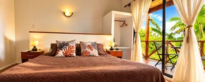 A bedroom with a large bed, two bedside tables, a nightstand, a wall-mounted light, and a view of a tropical garden through a sliding door. - Cabañas Te Maori (Hanga Roa)