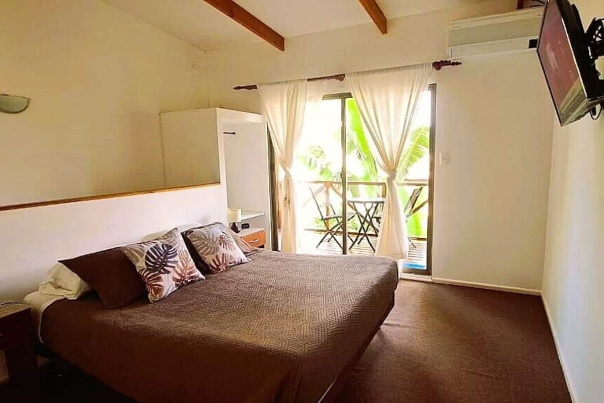 A hotel room with a bed, bedside table, lamp, and a view of the outside balcony.
