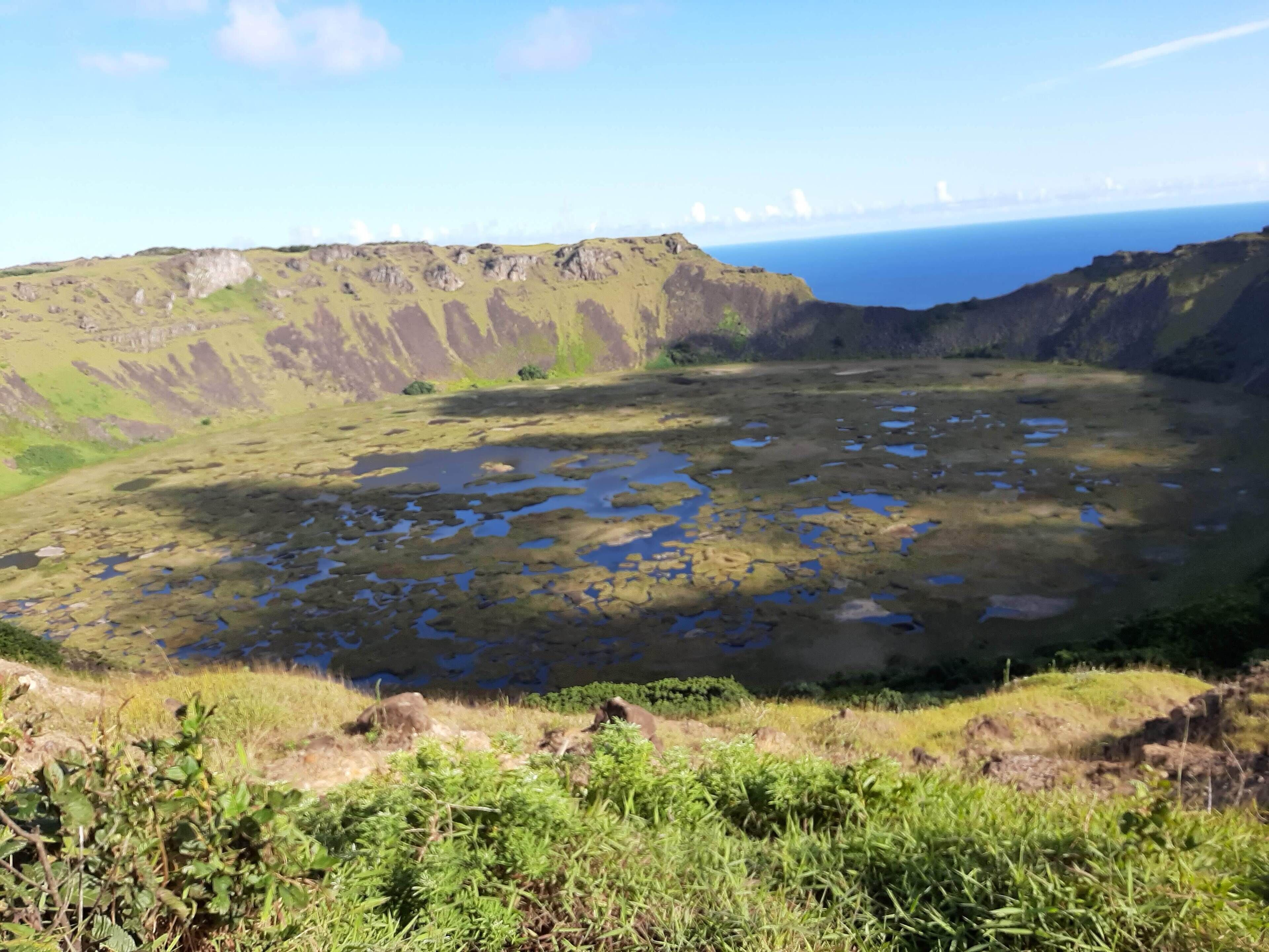 Un paysage naturel caractérisé par une dépression en forme de cratère, des zones herbeuses et une vue lointaine sur l’océan.