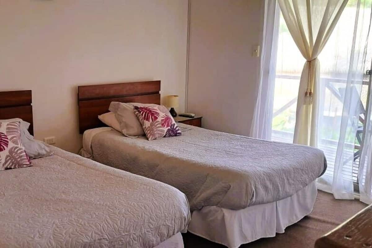 Two single beds with wooden headboards, a nightstand with a lamp, and a window with sheer curtains.