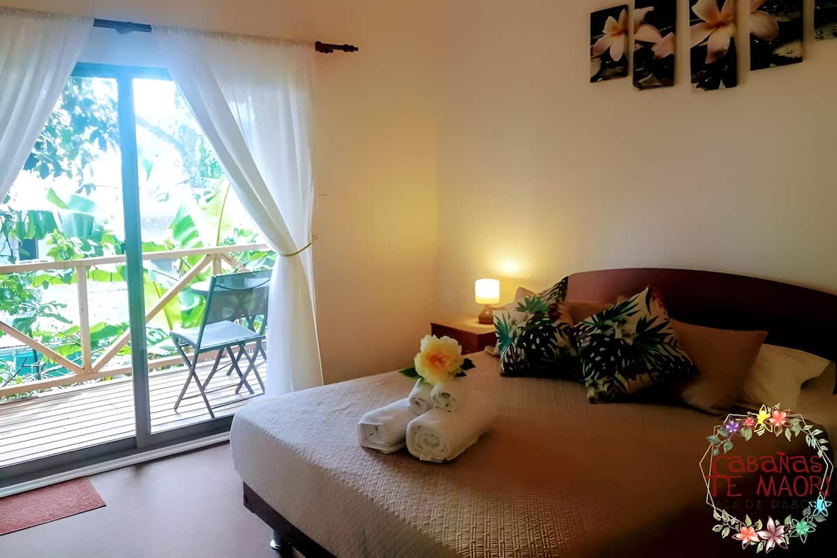A hotel room with a bed, a nightstand, a lamp, a window with curtains, and a view of greenery outside.