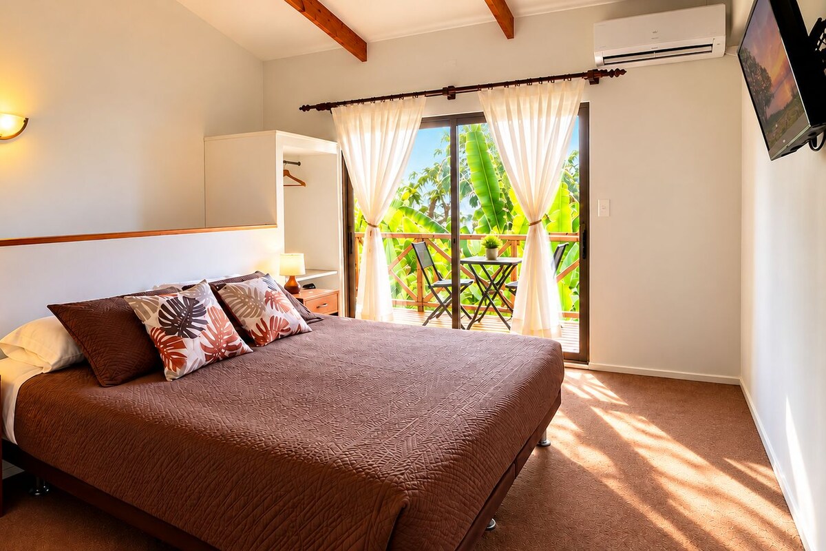 A bedroom with a bed, bedside table, lamp, and a view of a balcony with plants.