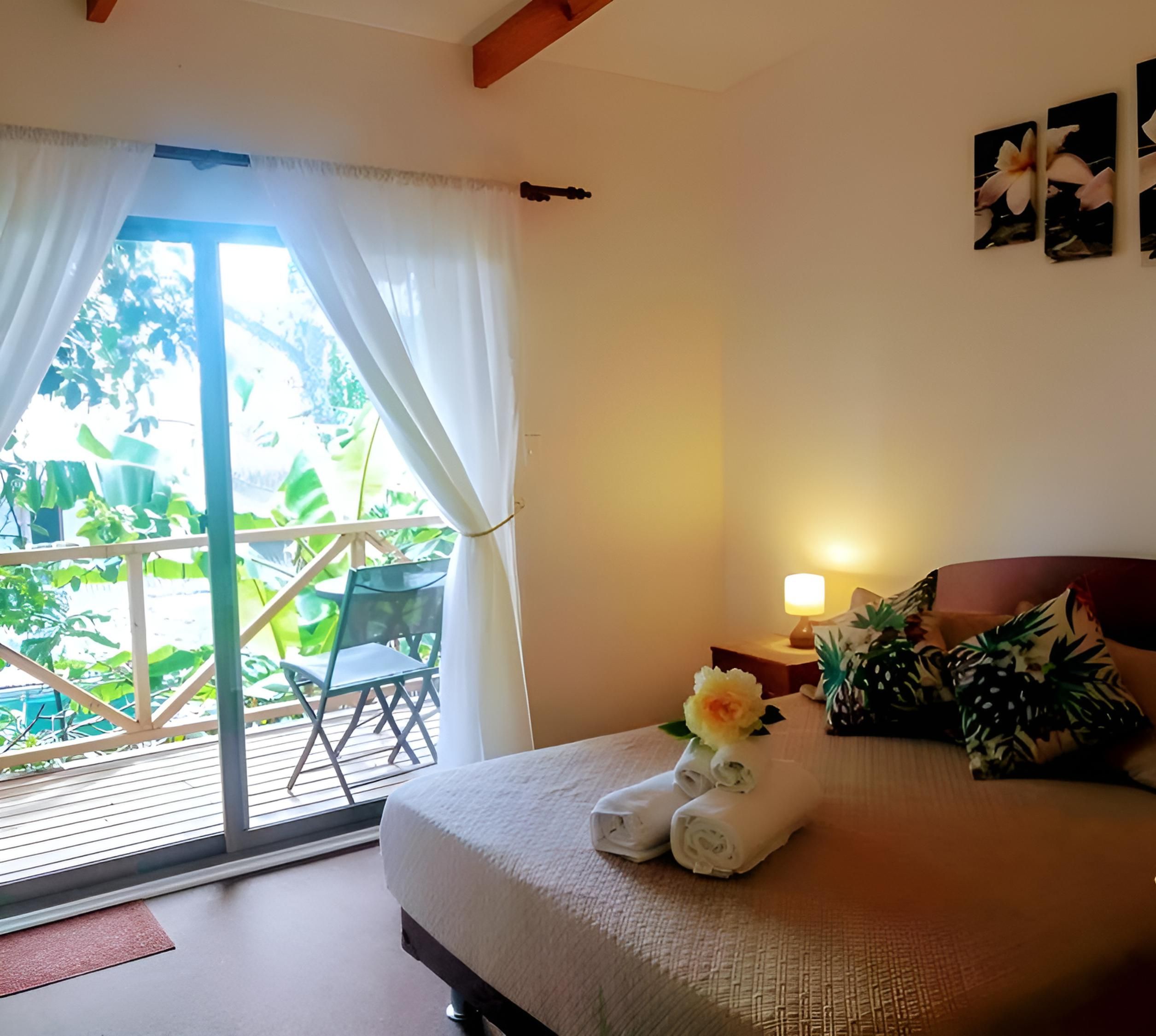 A hotel room with a bed, a nightstand, a lamp, a window with curtains, and a view of greenery outside.