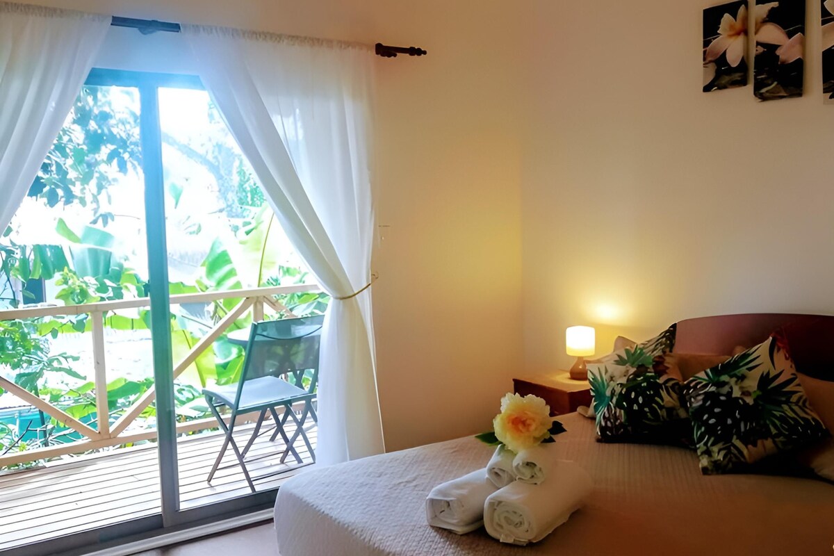 Habitación de hotel con cama, mesita de noche, lámpara, ventana con cortinas y vista a la vegetación.