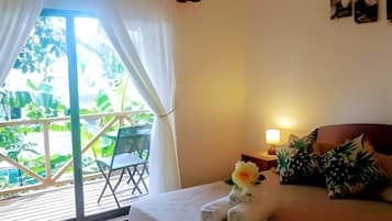 A hotel room with a bed, a nightstand, a lamp, a window with curtains, and a view of greenery outside.