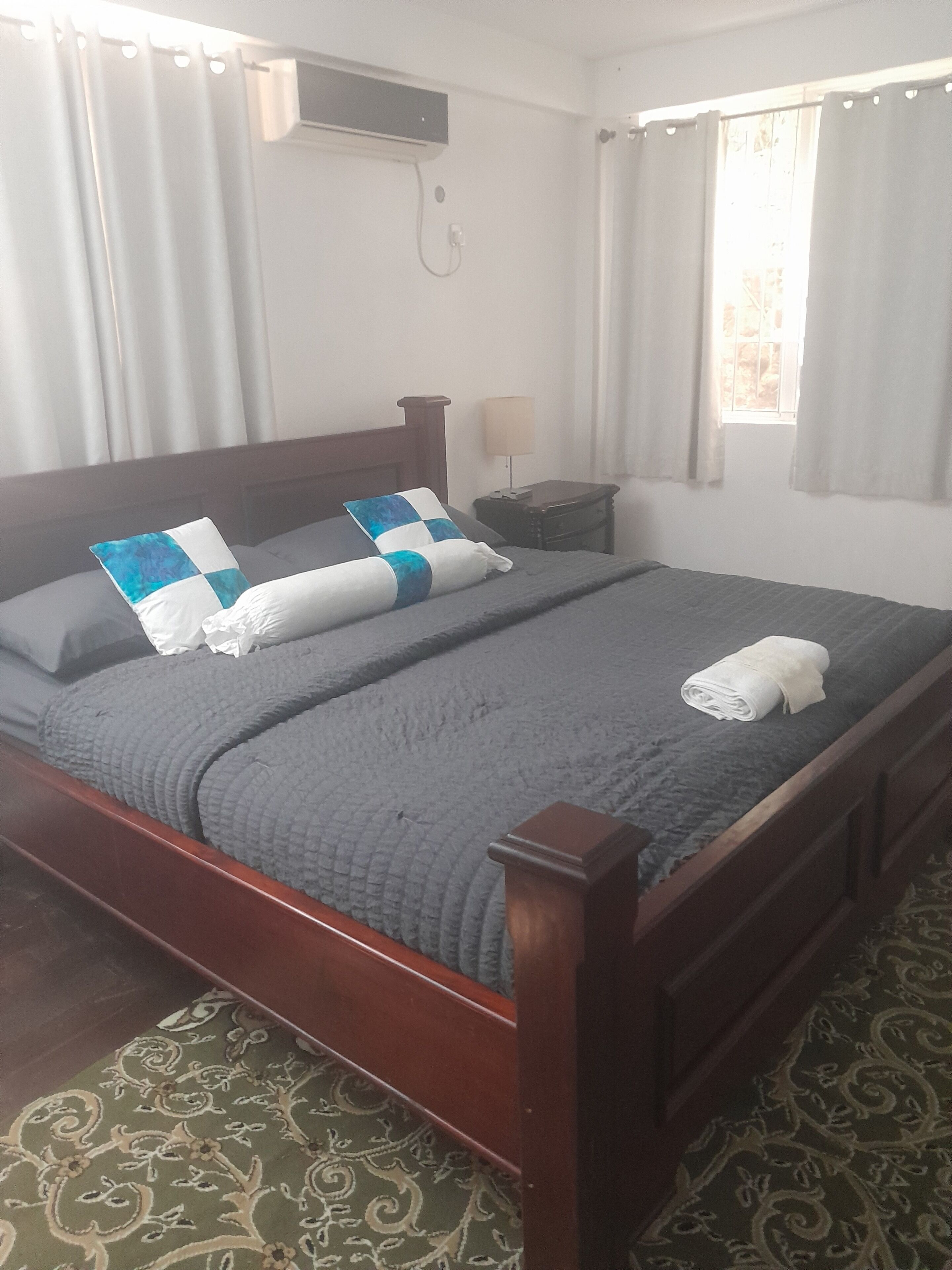 A neatly made bed with a grey quilt, two pillows, and a folded towel. There is a bedside table with a lamp, a wall-mounted air conditioning unit, and a window with curtains.