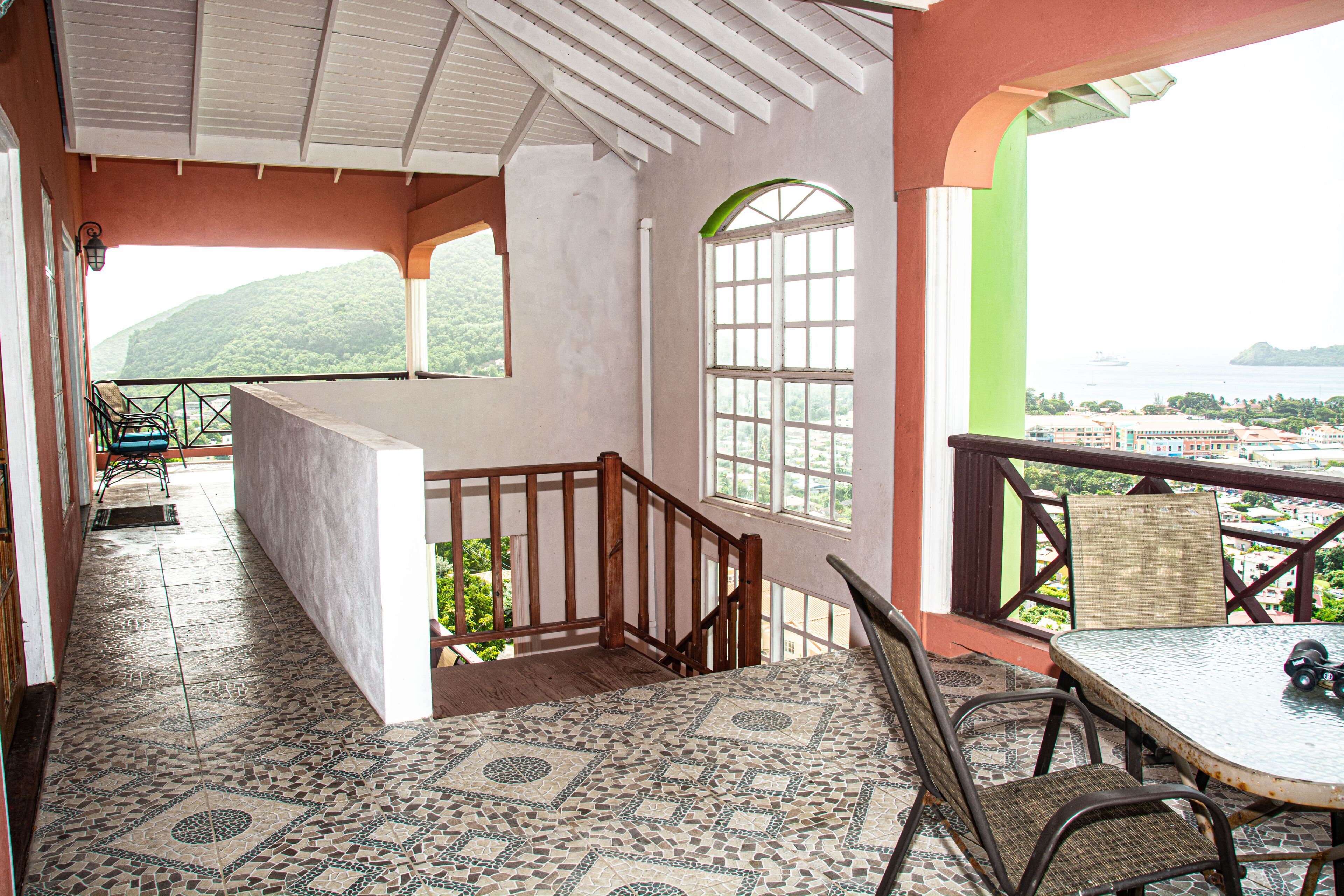 A spacious balcony with a view of the sea and hills, a tiled floor, and a table with chairs.