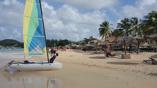 Een strand met een zeilboot, palmbomen en strandparasols.