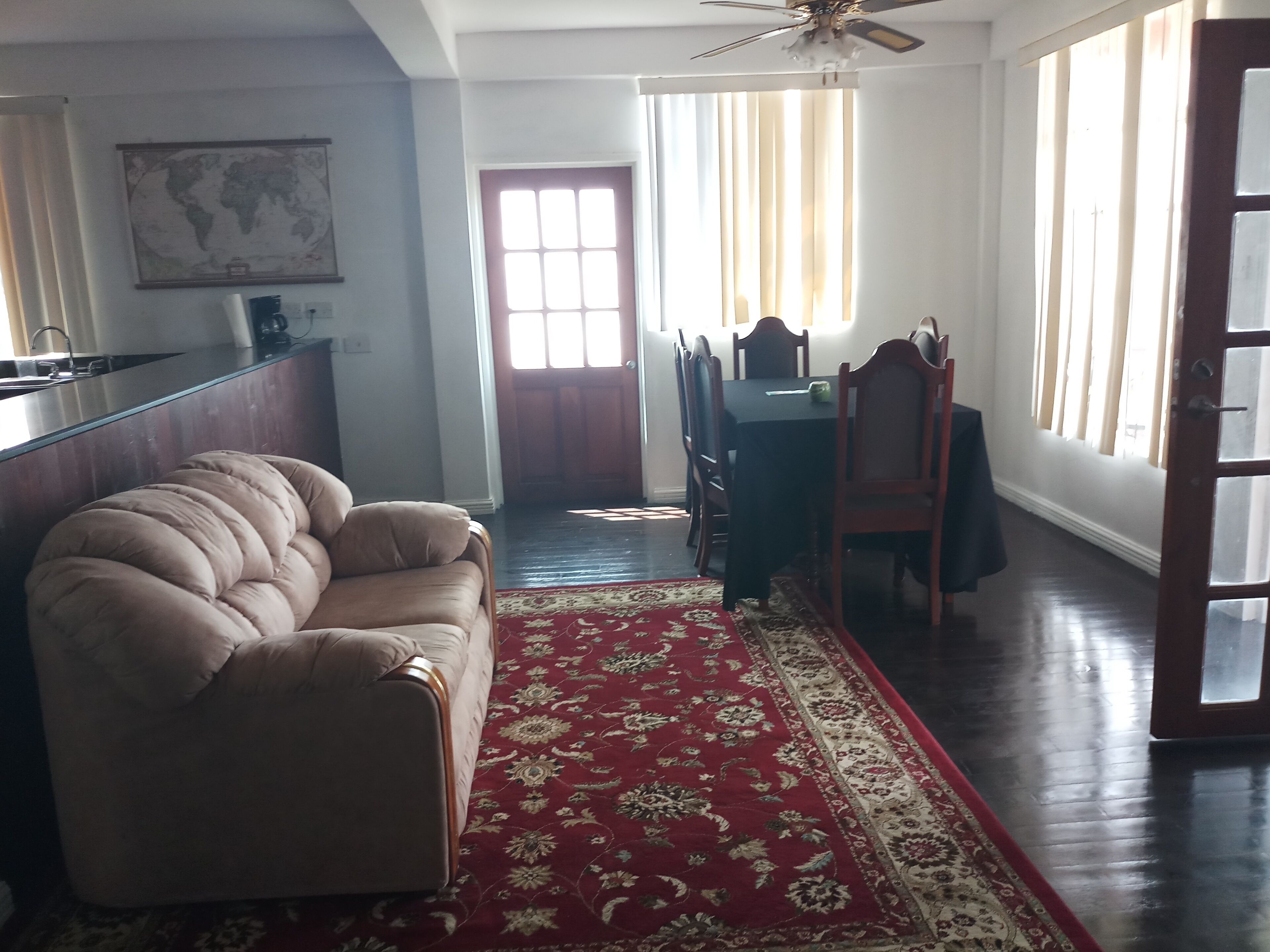 A living room with a beige sofa, a red patterned rug, a dining area with a table and chairs, and a kitchen area with a counter and cabinets.