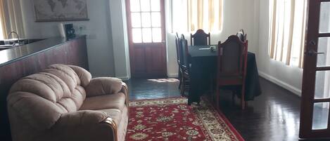 A living room with a beige sofa, a red patterned rug, a dining area with a table and chairs, and a kitchen area with a counter and cabinets.