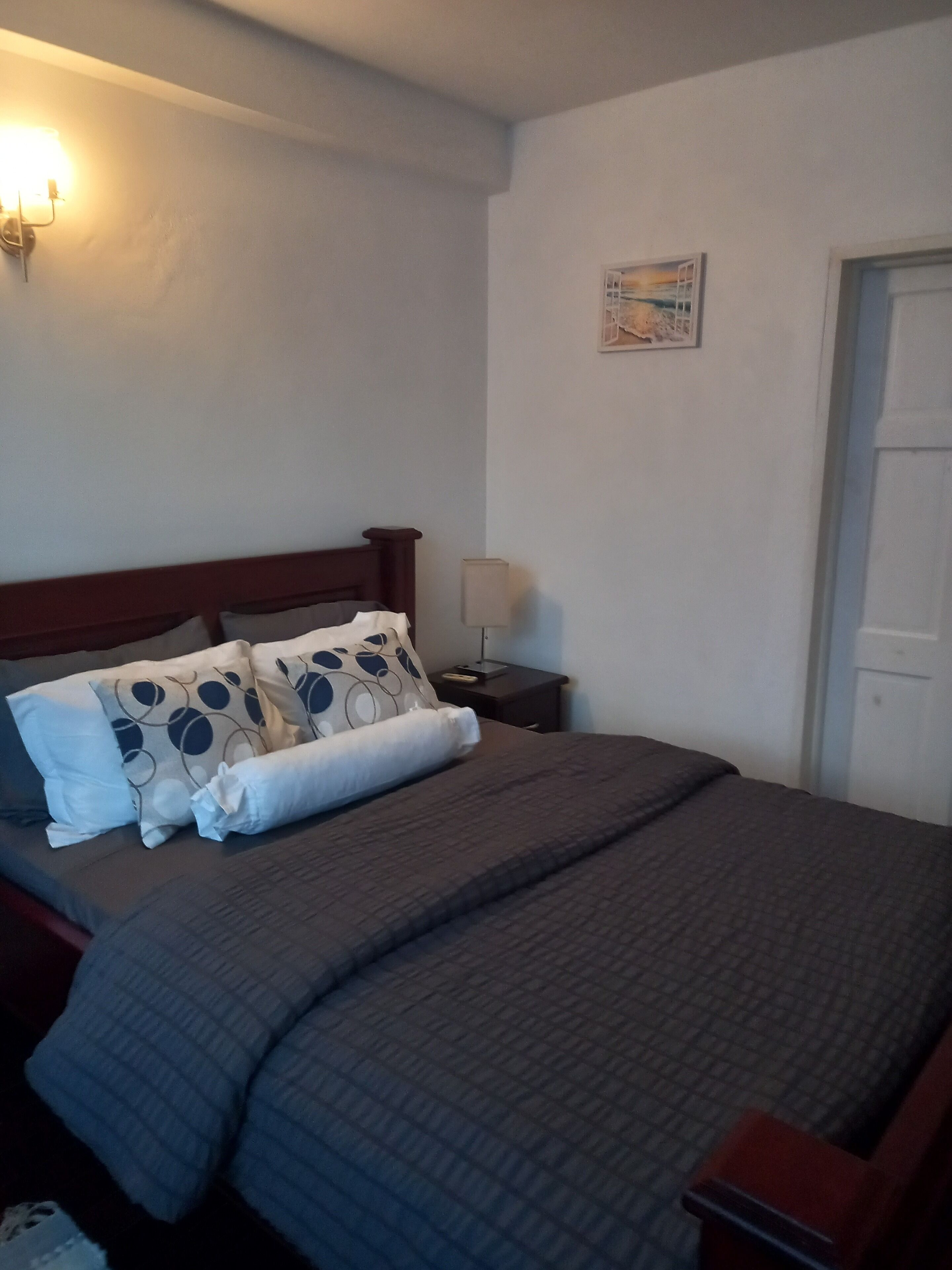 A neatly made bed with a grey quilt, a wooden headboard, and patterned pillows. There is a bedside lamp and a small table with a lamp on it. A framed picture hangs on the wall above the bed.