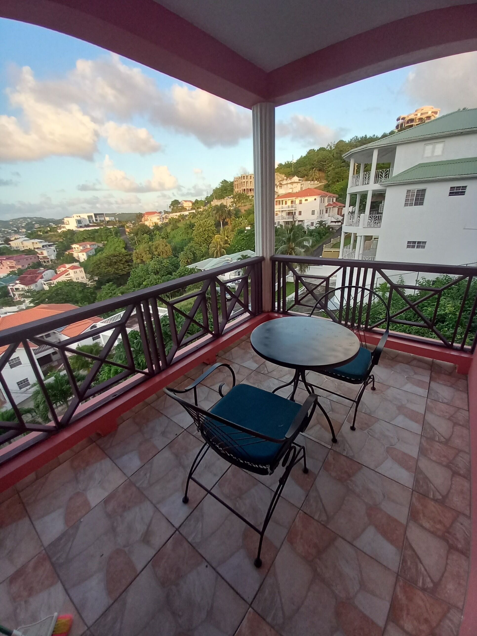 A balcony with a table and chairs, offering a view of a hillside with buildings and greenery.