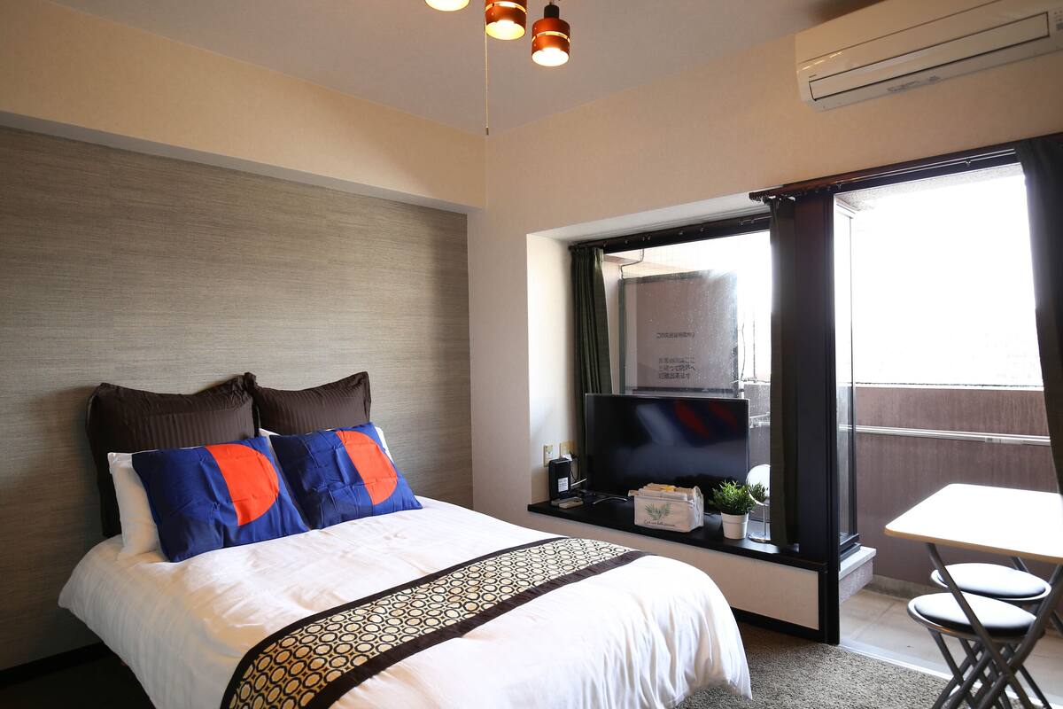 A hotel room with a bed, a TV, a small table with a plant, and a window with curtains.