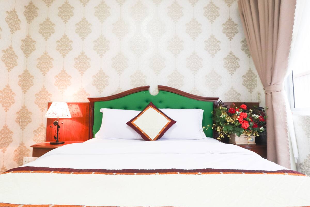 Una cama bien hecha con un cabecero verde, una almohada blanca y una almohada decorativa. Hay una mesita de noche con una lámpara y un jarrón con flores.