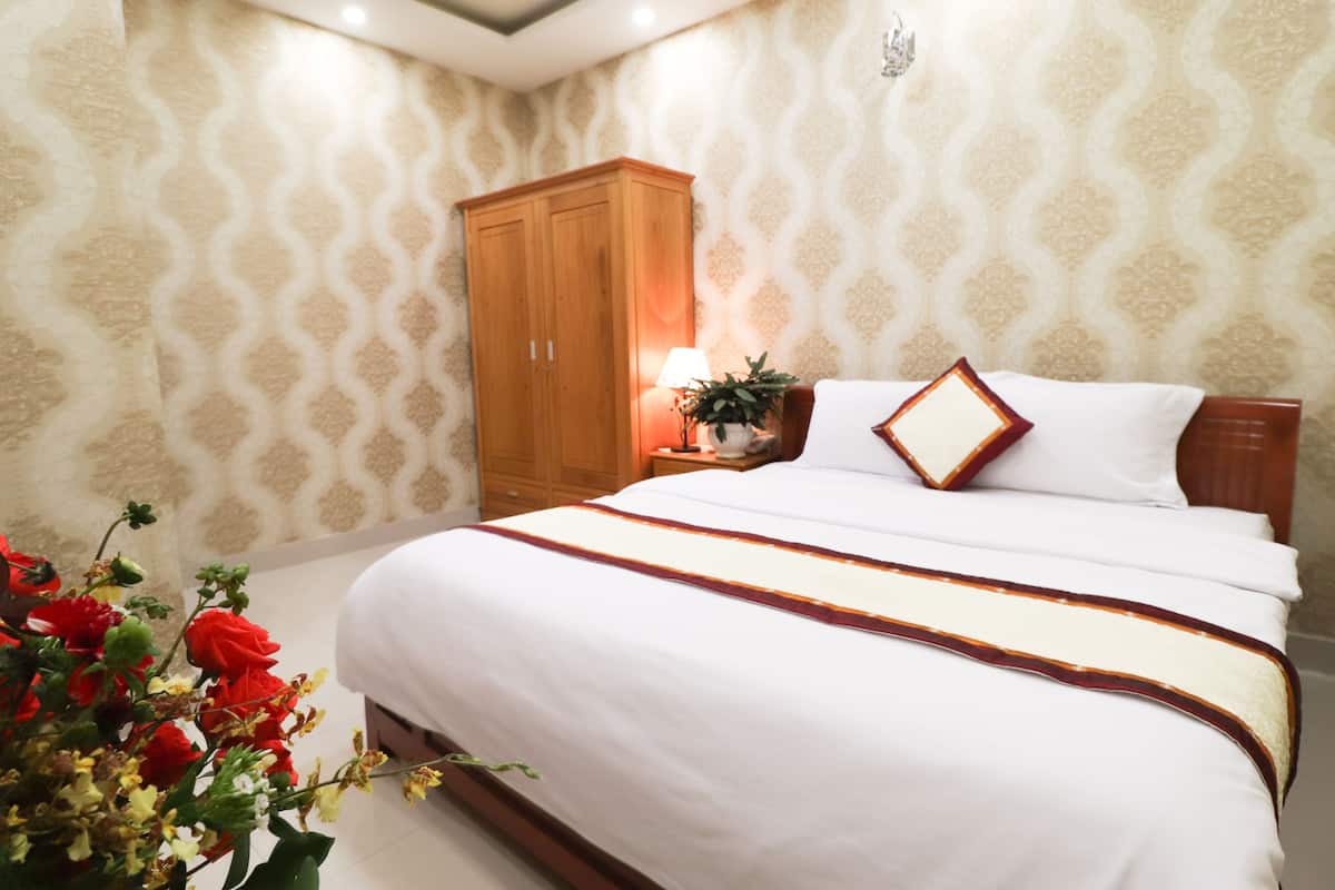 Un dormitorio ordenado con cama, un armario de madera, una mesita de noche con lámpara y un arreglo floral.