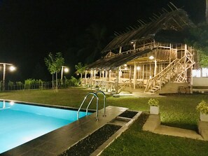 Piscina com deck de madeira, estrutura com telhado de palha e escadaria para área coberta.