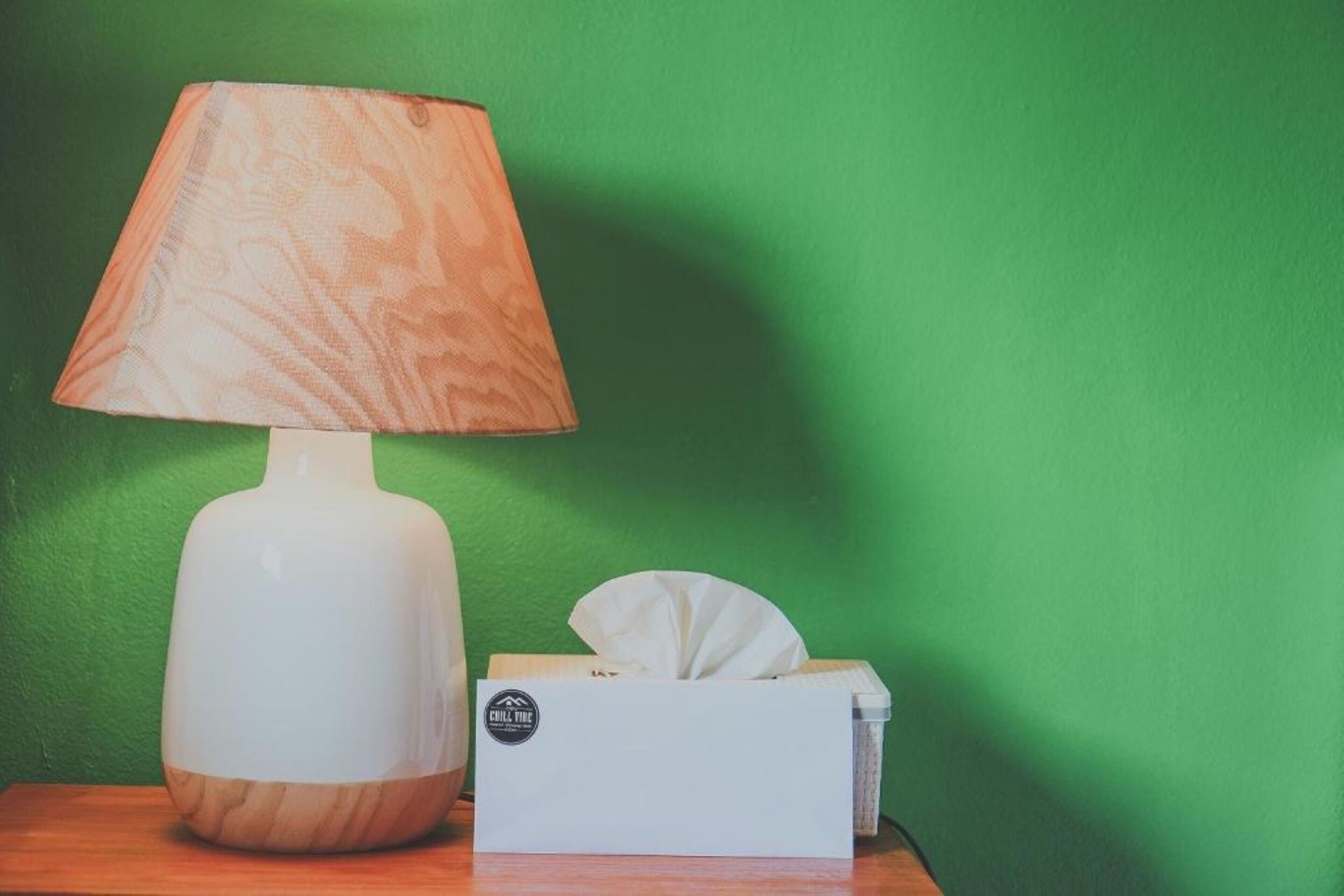 Una lámpara blanca con base de madera y una caja de pañuelos con etiqueta sobre una superficie de madera, frente a una pared verde.