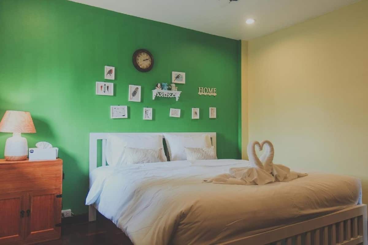 A bedroom with a green wall, a white bed, a wooden nightstand with a lamp, and wall decorations including a clock and framed pictures.