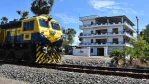 Um trem amarelo e azul sobre trilhos, com um edifício branco ao fundo.