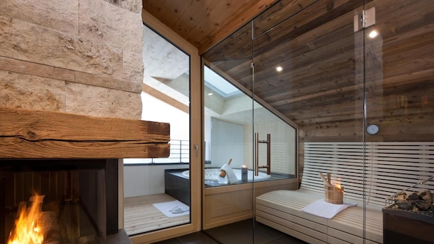 A modern spa room with a glass-enclosed sauna, a fireplace, and a wooden bench. - Hotel Walliserstube (Damüls)