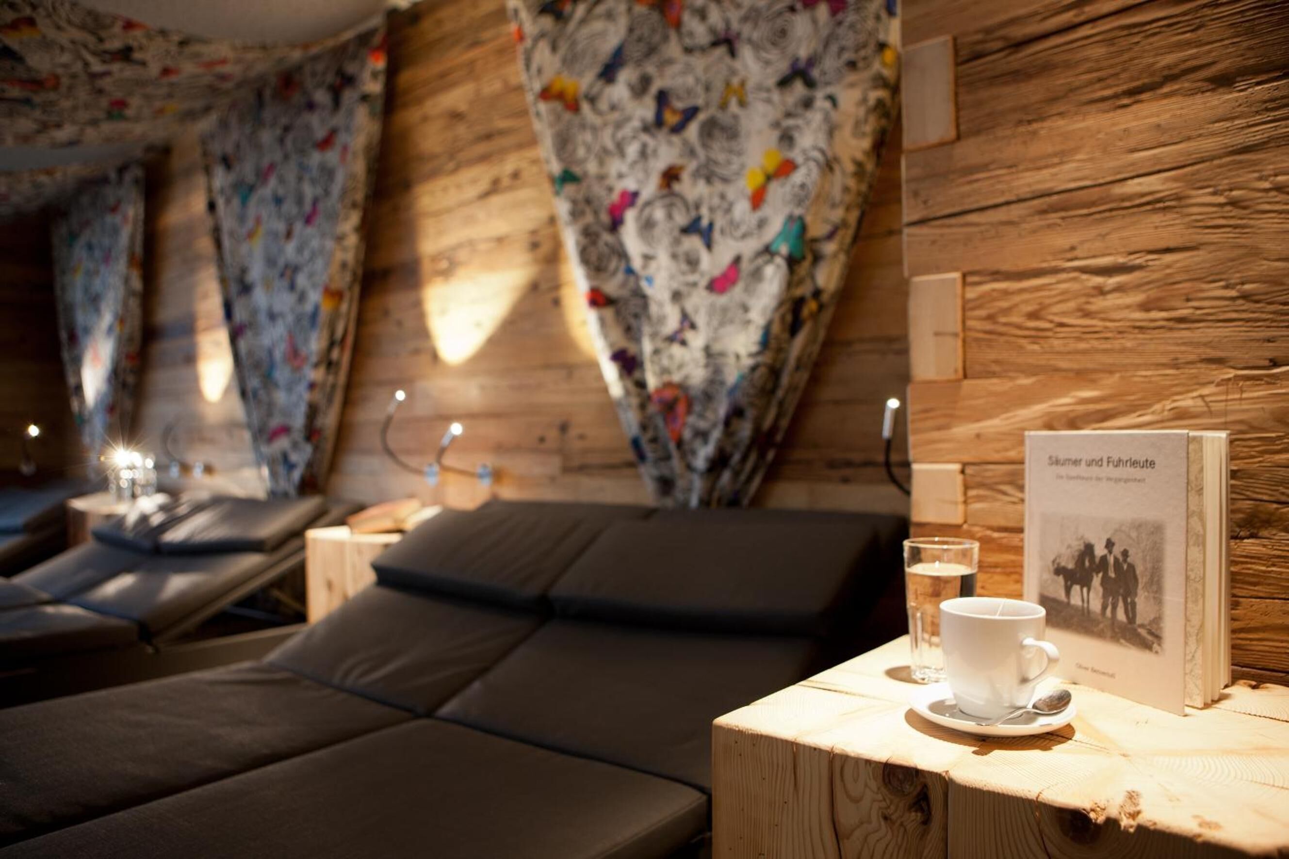 Ein gemütliches Zimmer mit einer Holzwand, einem Sofa, einem Tisch mit Tasse und Untertasse sowie einem Buch mit dem Titel 'Schulmeister und Fuhrleute'.