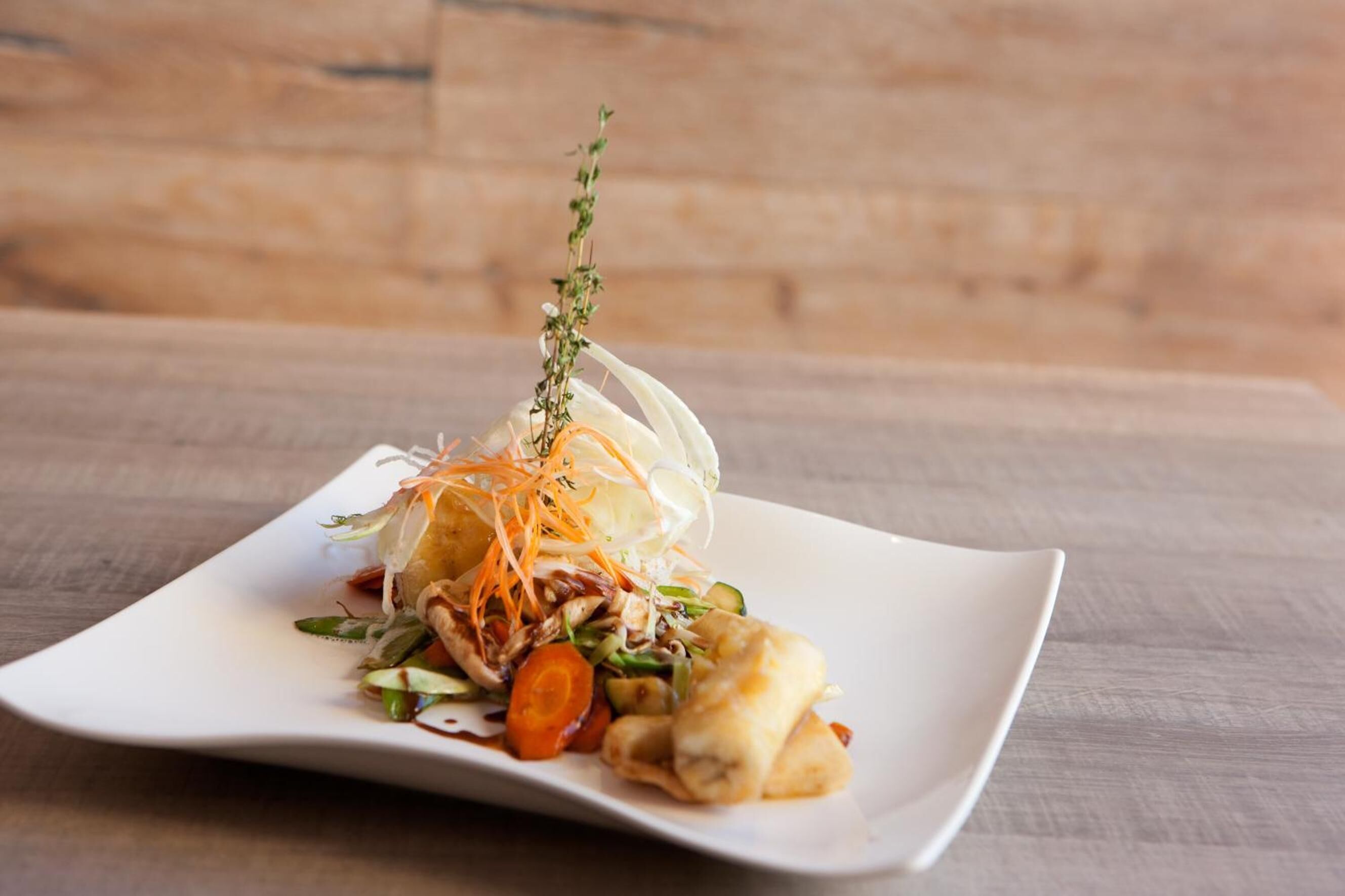 Une assiette carrée blanche contenant un plat composé de légumes et agrémenté d’une herbe décorative.