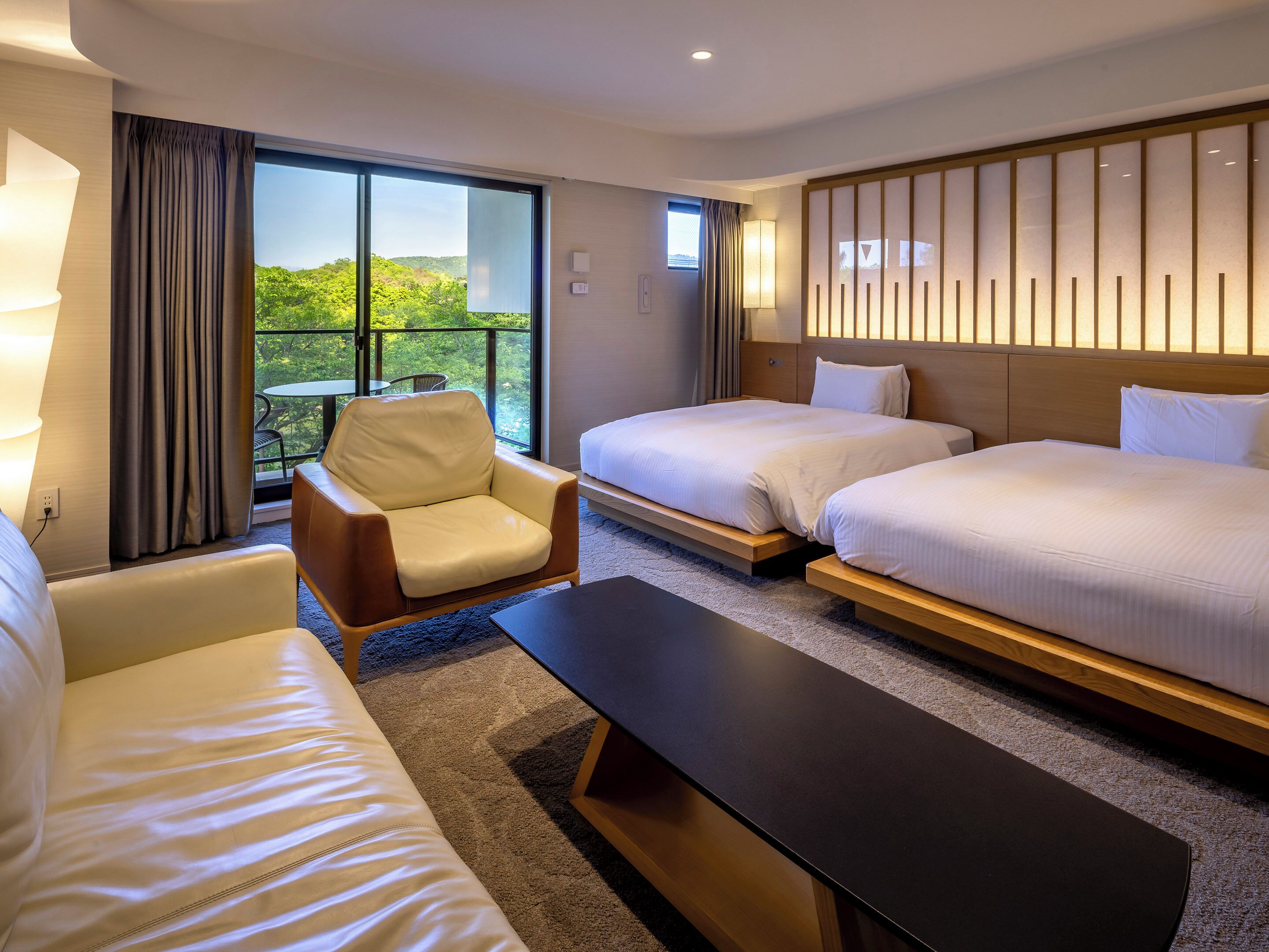 A hotel room with two beds, a sofa, a chair, a small table, and a balcony with a view of greenery.
