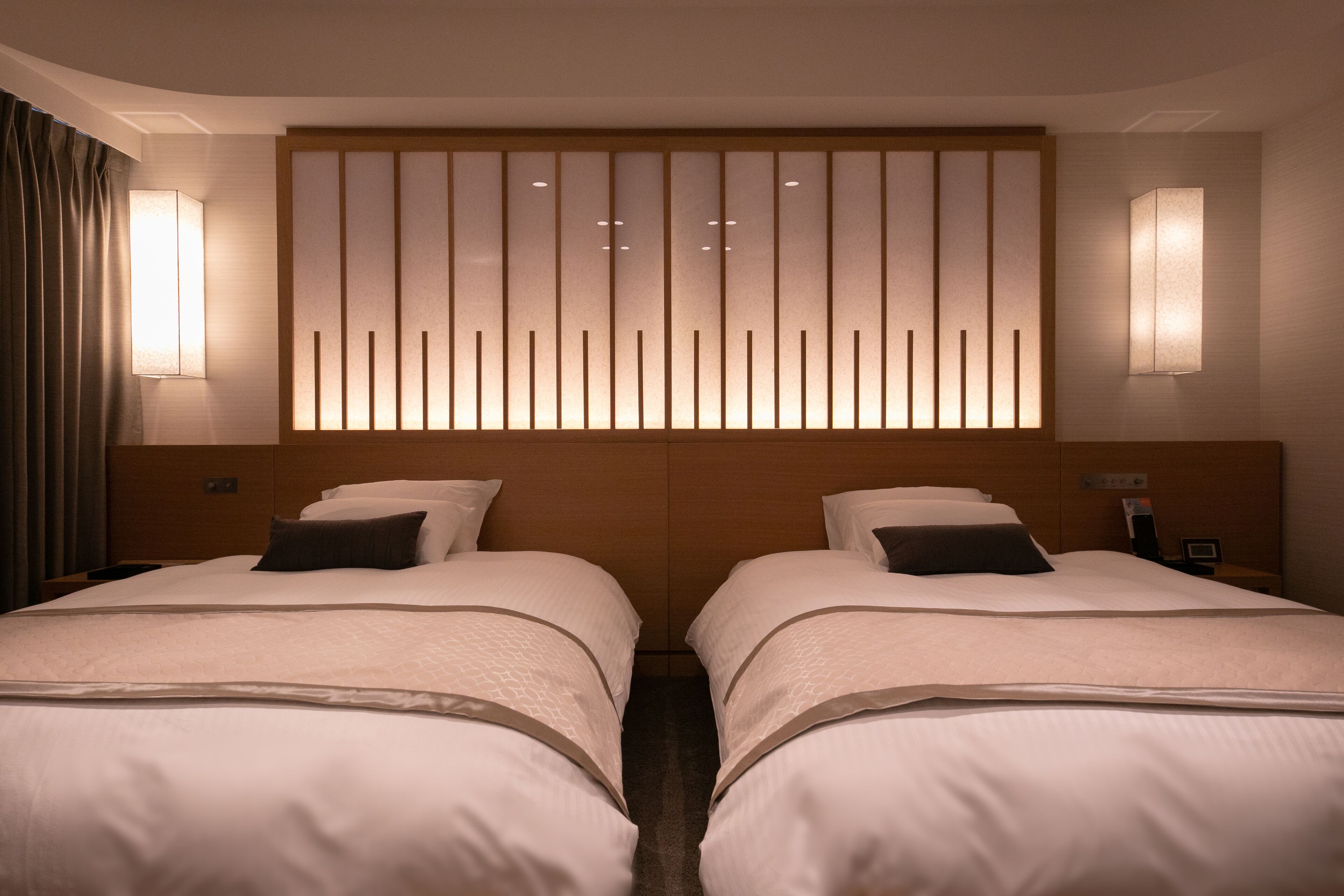 A hotel room with two beds, a wooden headboard with vertical slats, and wall-mounted lamps.