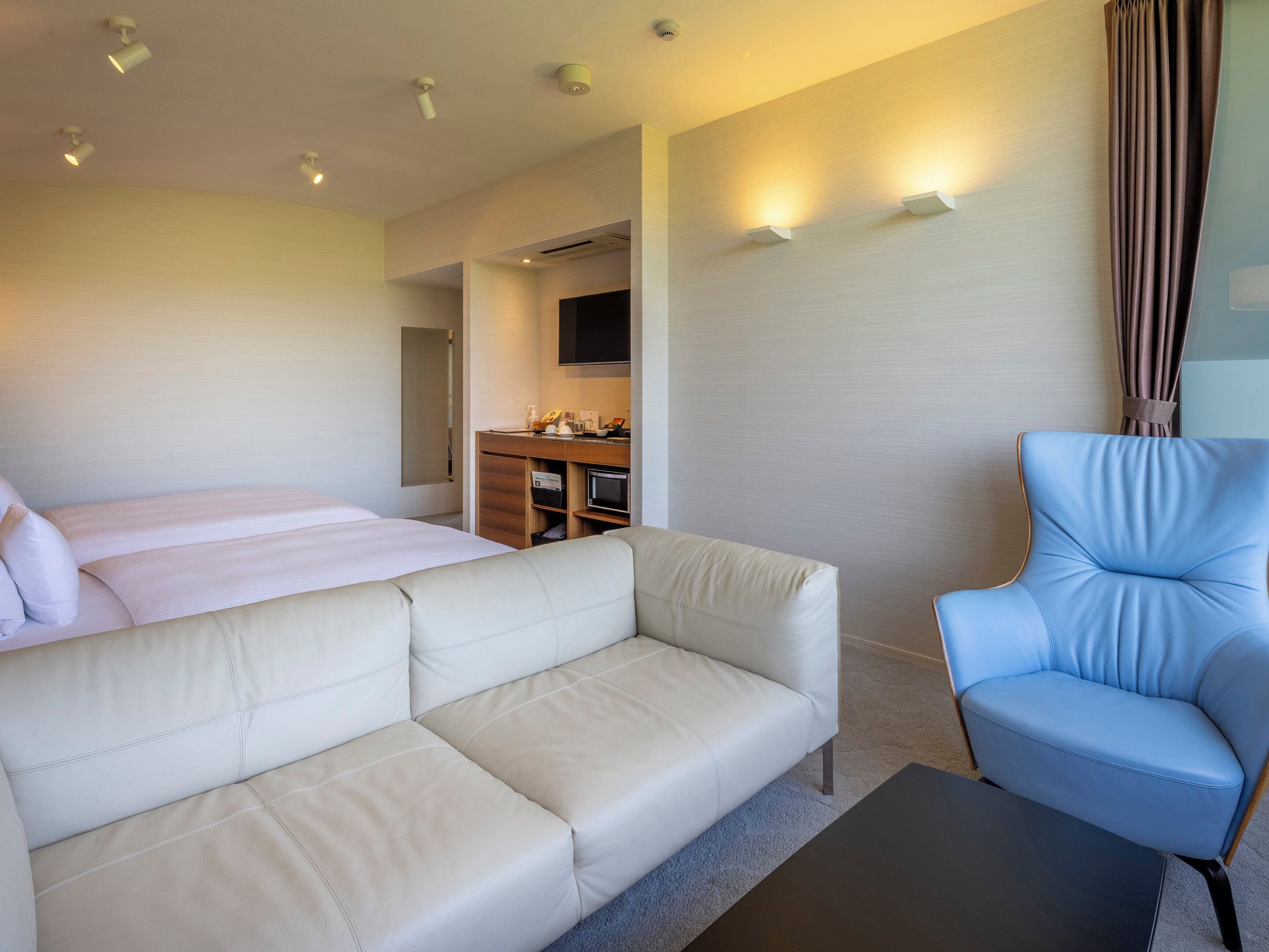 A hotel room with a bed, a sofa, a chair, a desk with a flat-screen TV, and recessed ceiling lights.