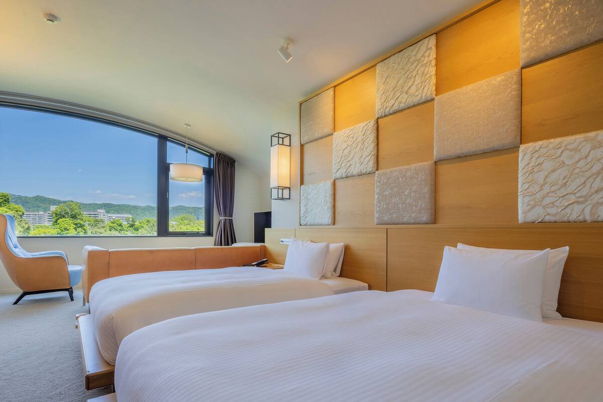 A hotel room with two beds, a large window with a view of greenery, and a modern design with a textured wall panel.