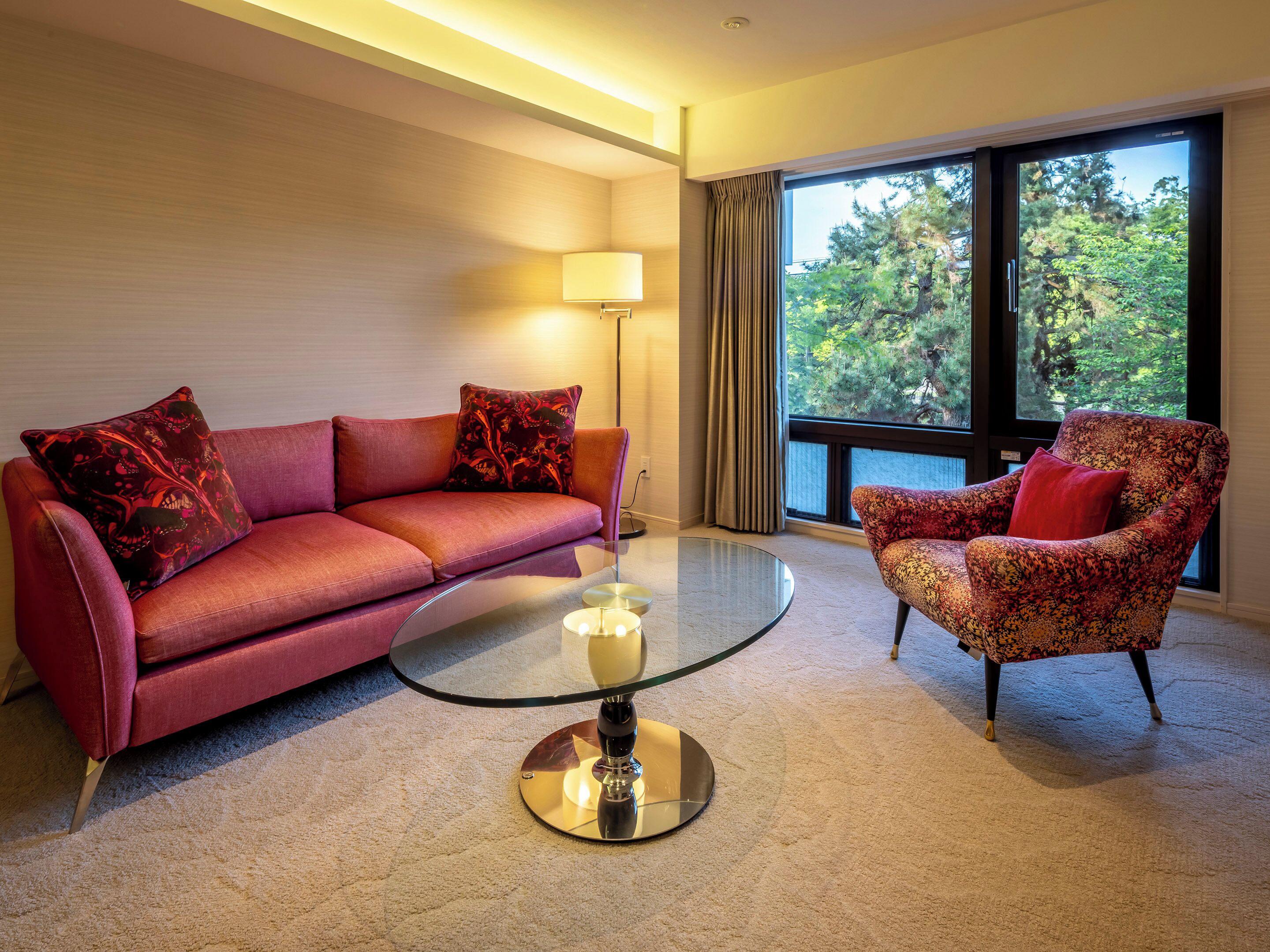 Habitación de hotel moderna con un sofá rojo, un sillón estampado, una mesa de centro de vidrio y un ventanal con vistas a árboles.