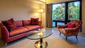 Habitación de hotel moderna con un sofá rojo, un sillón estampado, una mesa de centro de vidrio y un ventanal con vistas a árboles.
