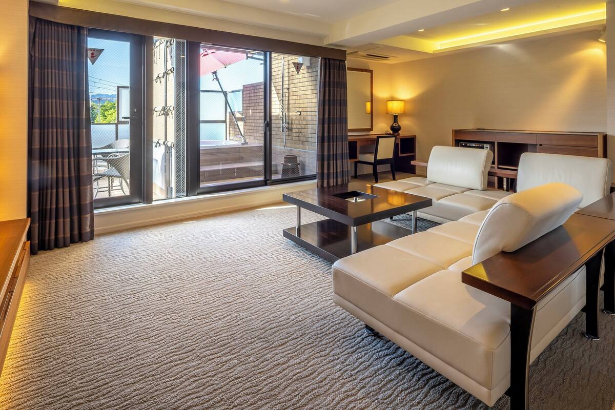 A modern hotel room with a large sliding glass door leading to a balcony, a sofa, a small table, and a desk with a lamp.