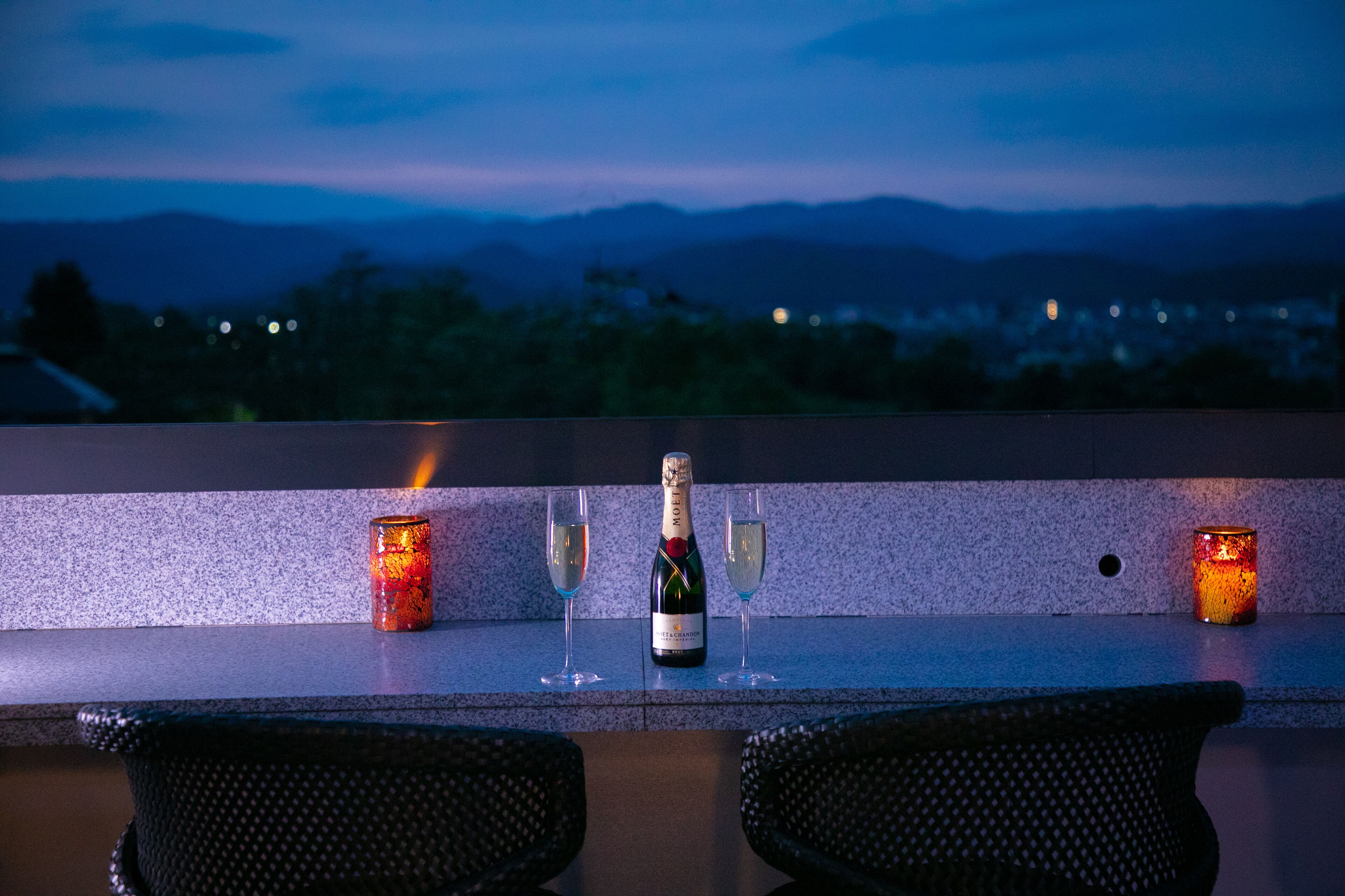 A bar setting with a bottle of champagne, two glasses, and two candles on a counter with a view of mountains in the background.