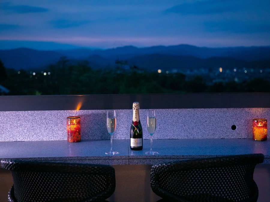A bar setting with a bottle of champagne, two glasses, and two candles on a counter with a view of mountains in the background.
