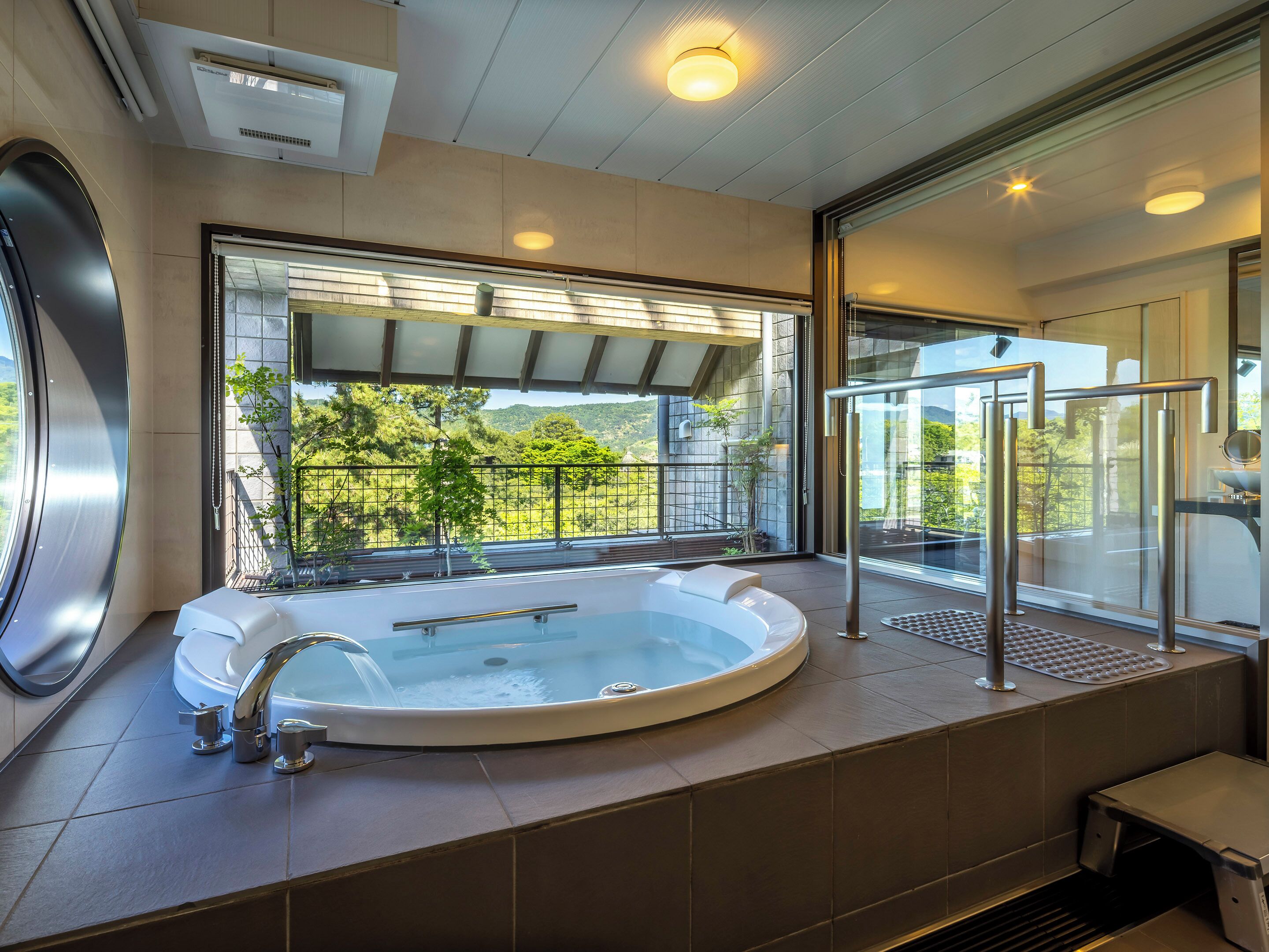 A modern bathroom with a large bathtub, a glass-enclosed shower area, and a view of greenery outside.