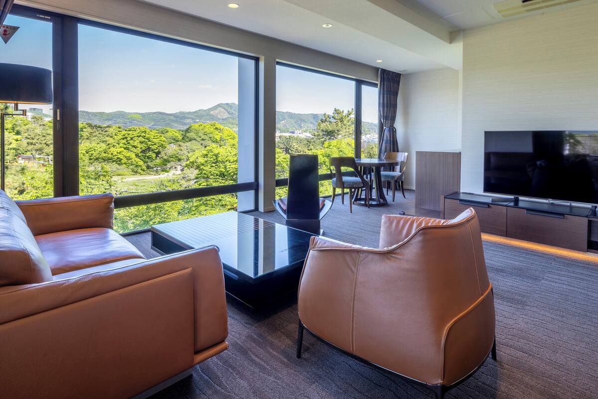 A modern hotel room with a large window offering a scenic view of green hills. There is a leather sofa, a coffee table, and a TV in the room.