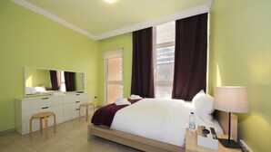 Een hotelkamer met een bed, nachtkastje met lamp, spiegel, dressoir en raam met gordijnen.