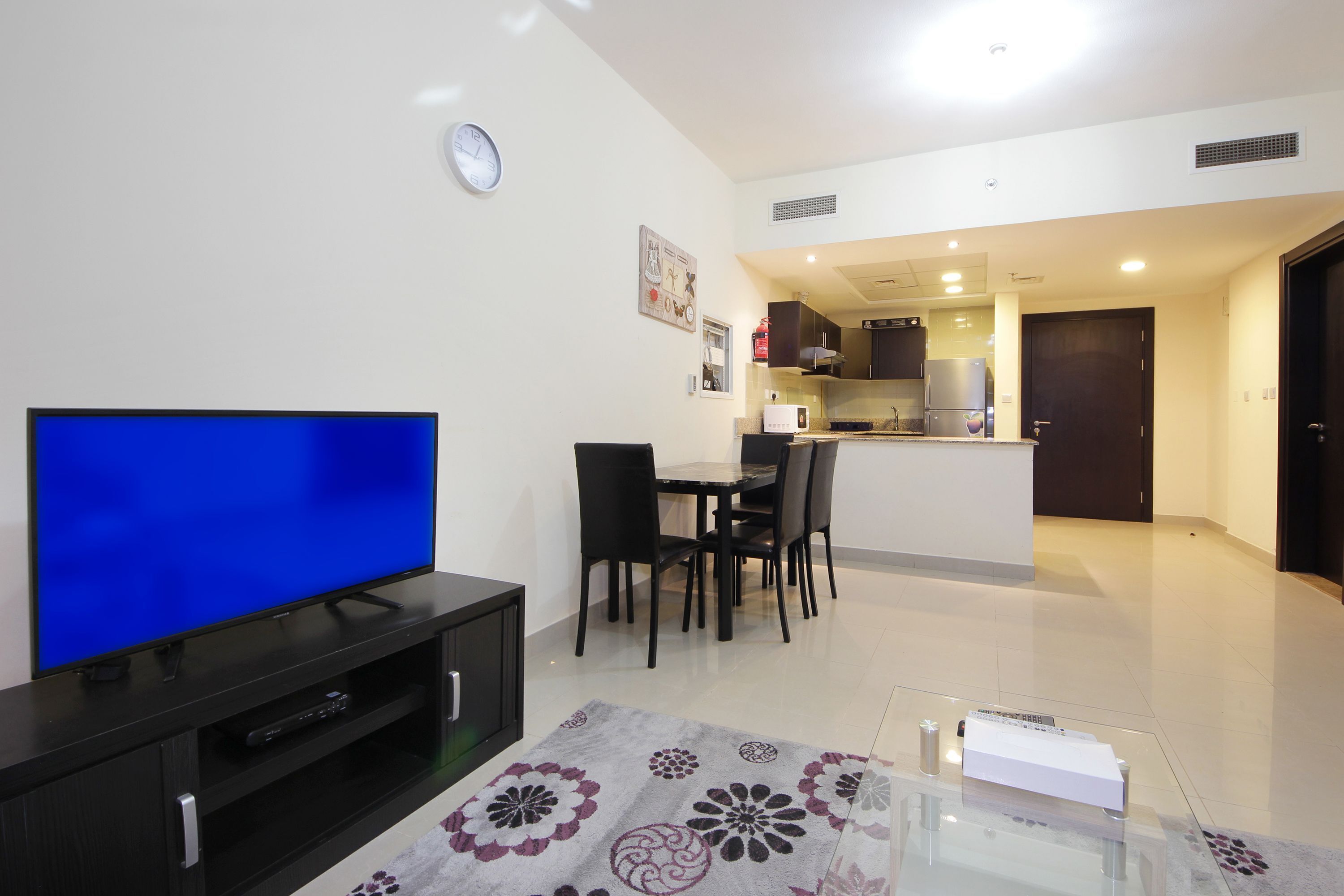 A modern living room with a flat-screen TV, a dining area with a table and chairs, and a kitchen in the background.