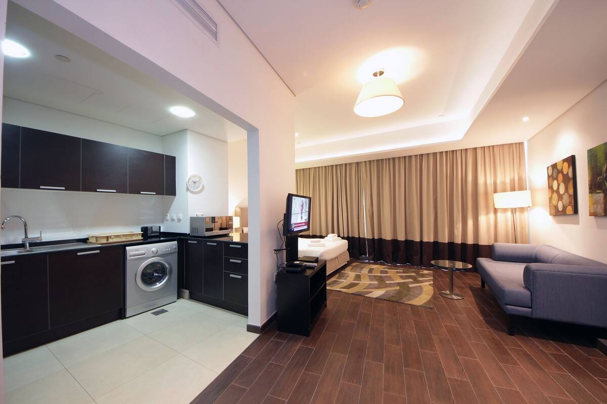 A modern hotel room with a kitchen area, a living room with a sofa and a TV, and a large window with curtains.