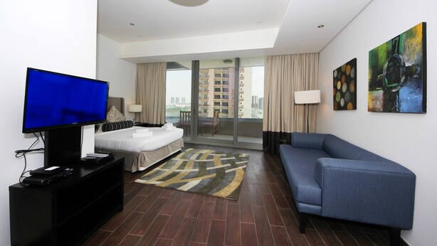 A modern hotel room with a bed, a TV, a sofa, and a balcony with a city view. - Signature Holiday Home - Matrix Tower (Dubai)