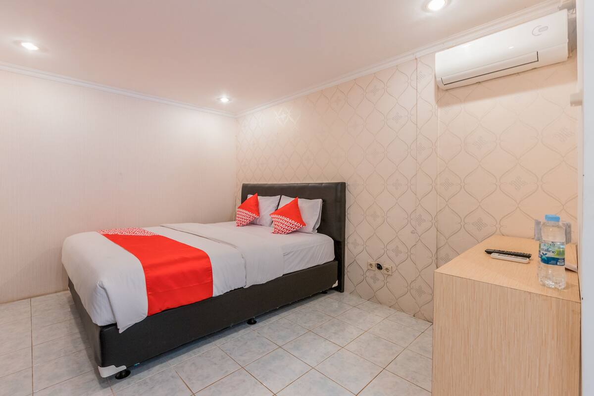 A neatly made bed with a red and white bedspread, a wooden bedside table with a remote control and a bottle of water, and a wall-mounted air conditioner.