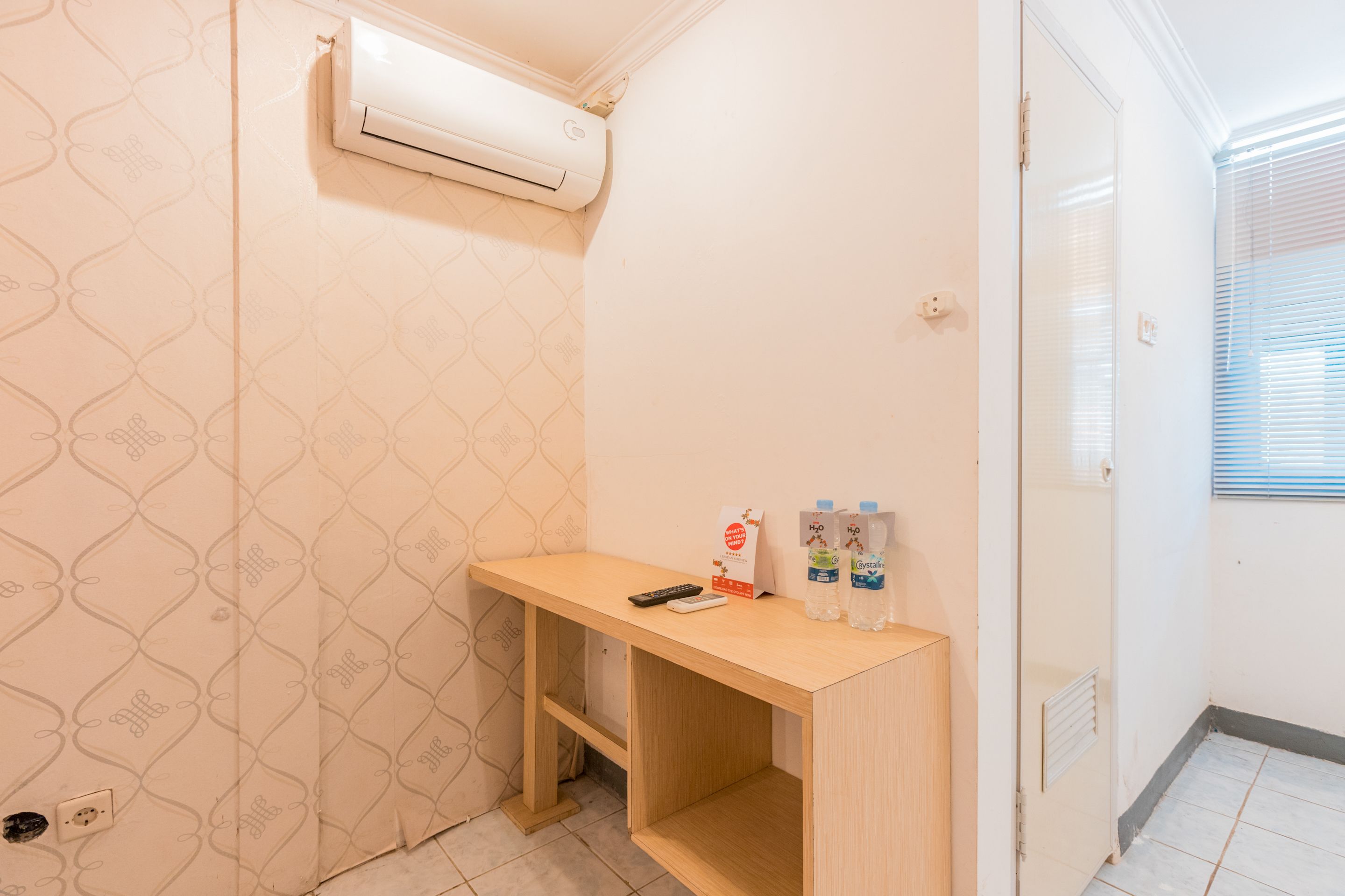 A small room with a wooden desk, a wall-mounted air conditioner, a door, and a window with blinds.
